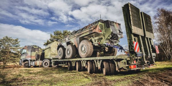 Military heavy load transporter with camouflaged armored vehicle on low loader in a forest landscape.